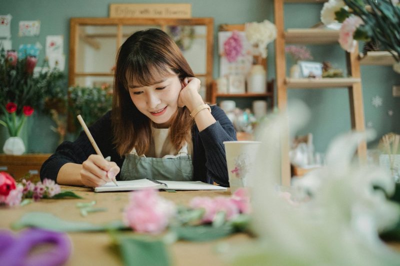 Smiling young florist in an apron writing notes in a cozy, colorful flower shop.