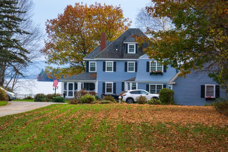 A blue house nestled among vibrant autumn leaves beside a tranquil lake in a rural setting.