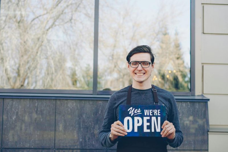 Smiling young business owner in glasses holding a 'Yes, we're open' sign outside their store.