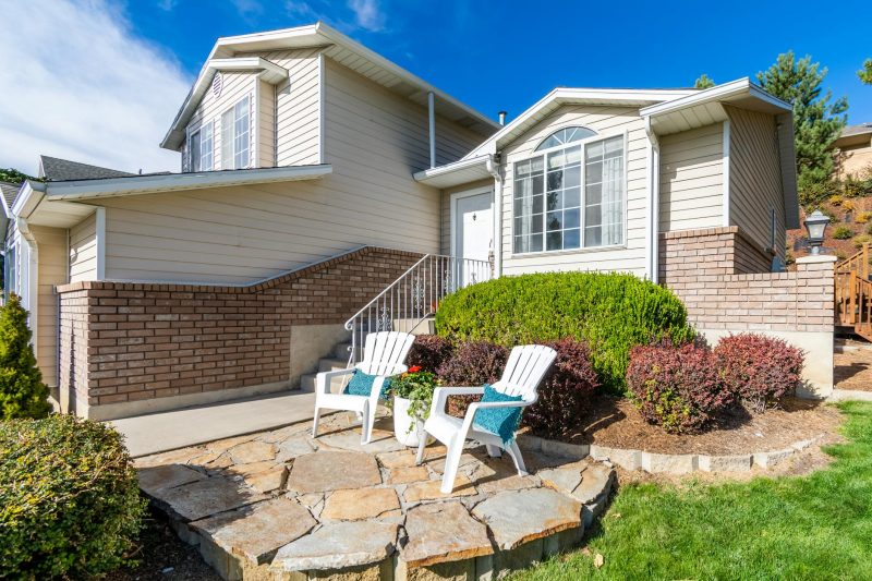 Elegant suburban house with a porch, seating area, and well-maintained landscaping under bright daylight.