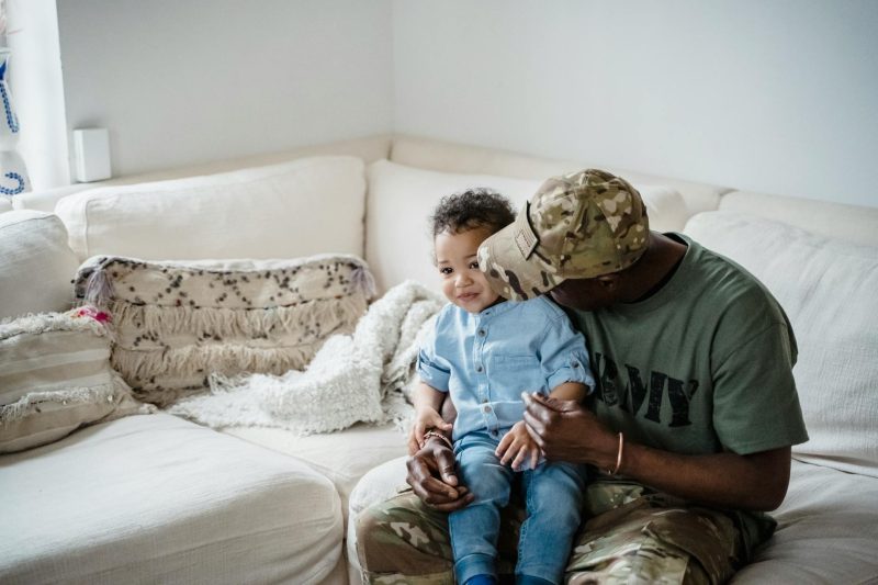 A father in army attire embraces his child on a cozy couch at home.