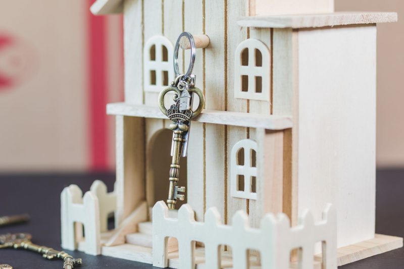 Miniature wooden house next to a vintage key on a light surface.